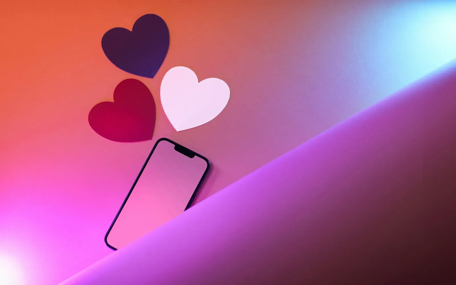 Smartphone mit drei Papierherzen auf pink-violettem Hintergrund – Tinder oder Lovoo im Vergleich